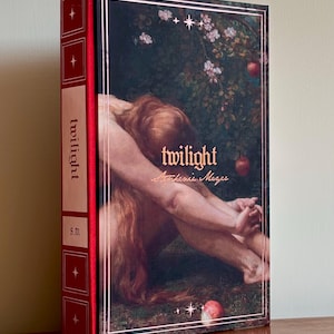 Puede incluir: Un libro de tapa dura titulado Twilight de Stephenie Meyer con un lomo rojo que presenta paneles blancos y detalles de estrellas doradas. La portada muestra una pintura clásica de una figura con largo cabello rojo entre manzanos.