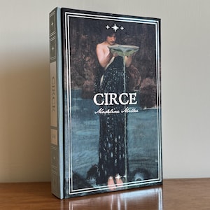 Op de afbeelding: Een hardcover boek getiteld Circe door Madeline Miller met een donkere, geïllustreerde kaft waarop een figuur in een zwarte jurk vloeistof uit een kom giet. Het boek heeft een lichtblauwe rug met de titel en initialen in witte tekst.