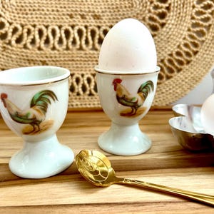 May include: Two white porcelain egg cups featuring a colorful rooster illustration with green, brown, and red details. One cup holds a white egg. A gold-toned metal spoon with a decorative handle rests in front of the cups on a wooden surface.