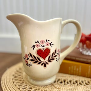 May include: A cream-colored ceramic pitcher featuring a central design of a red heart with black polka dots, surrounded by dark brown leafy branches and small pink flowers. The pitcher has a curved handle and a wide pouring spout.