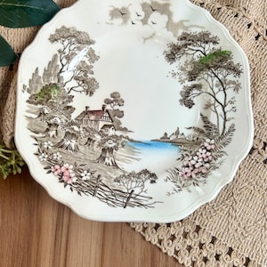 May include: A cream-colored ceramic plate with a scalloped edge featuring a brown transferware landscape scene of a house, trees, and wheat sheaves, accented with hand-painted touches of blue water and pink flowers.