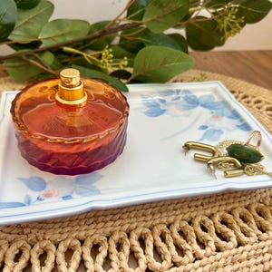 Puede incluir: Una bandeja rectangular de cerámica blanca con un patrón floral azul, que contiene un frasco de perfume redondo de vidrio ámbar con un atomizador dorado y un collar de cadena dorada con un colgante de piedra verde oscuro y pendientes dorados.