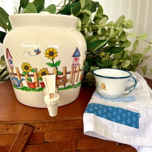 Pode incluir: Um dispensador de bebidas de cerâmica na cor creme com uma cena de jardim colorida apresentando girassóis, uma cerca de madeira, casas de pássaros e uma abelha. Uma torneira de plástico branca está fixada na frente. Ao lado, há uma pequena xícara branca com borda azul.