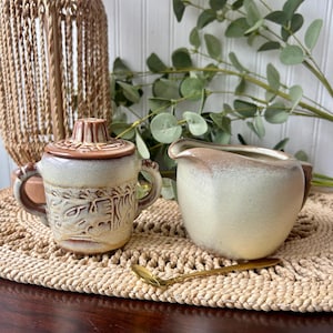 May include: A ceramic sugar bowl with a lid and two handles, a matching creamer pitcher, and a small gold-toned metal spoon. Both ceramic pieces feature a gradient finish transitioning from a creamy off-white base to a warm brown rim.