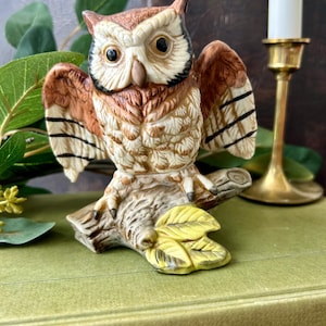 May include: A ceramic owl figurine with brown, white, and black plumage, perched on a textured branch with yellow leaves. The owl has its wings partially spread, dark eyes, and a light-colored beak, showcasing detailed feather textures.
