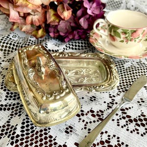 May include: A silver-plated butter dish with an ornate, embossed border and a central handle on the lid, accompanied by a matching silver-toned butter knife. The rectangular base features a decorative floral pattern etched into the bottom surface.