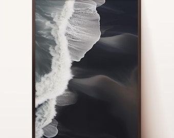 May include: An aerial photograph of a wave crashing on a black sand beach. The white foam of the wave contrasts sharply with the dark sand.