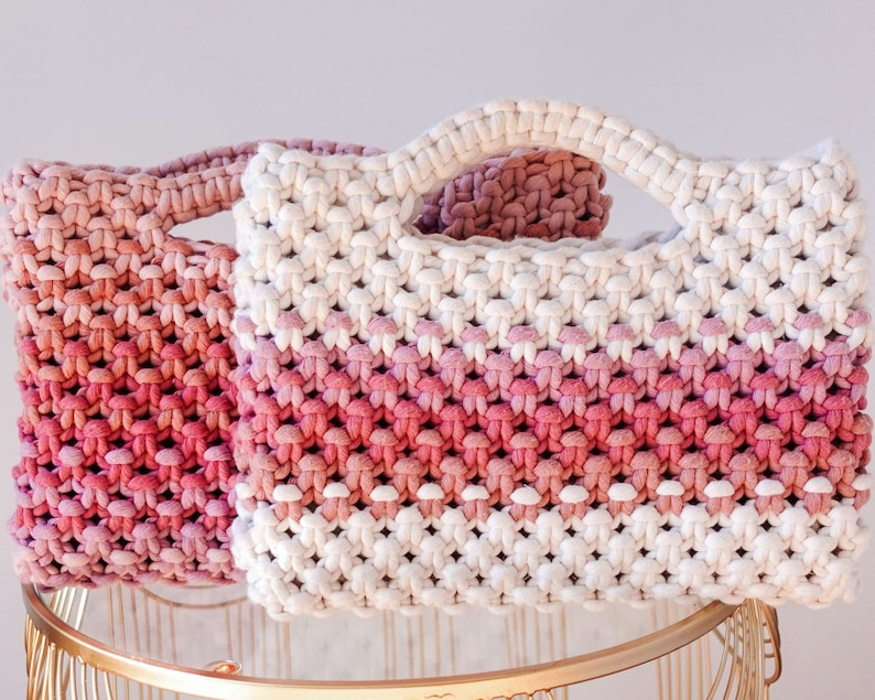 May include: Two macrame handbags in shades of pink and white. The bags have a rectangular shape with a handle. The macrame design creates a textured appearance. One bag is a solid pink, while the other transitions from white to pink.