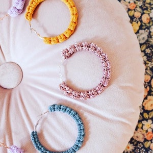 May include: Four crocheted hoop earrings in different colors: purple, yellow, pink, and blue. The earrings are displayed on a pink velvet surface.