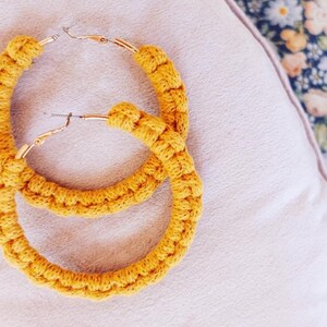 May include: A pair of large, yellow crocheted hoop earrings with gold metal hoops. The earrings are on a light pink fabric background.