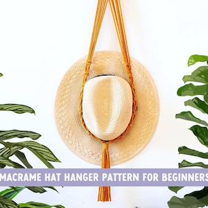 May include: A macrame hat hanger with a natural straw hat hanging from it. The hanger is made with a thick, natural-coloured cord and has a tassel at the bottom. The hat hanger is mounted on a white wall.