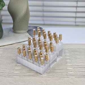 Peut inclure: Un organisateur en plastique transparent rempli de petits flacons en verre surmontés de bouchons dorés. Les flacons sont disposés en grille, créant une présentation soignée et organisée. L'arrière-plan comprend un vase décoratif et un tissu rayé.