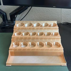 May include: A light brown, tiered paint bottle holder with three rows of circular openings and a bottom tray. The holder is made of a light-colored material and is designed to hold multiple paint bottles.