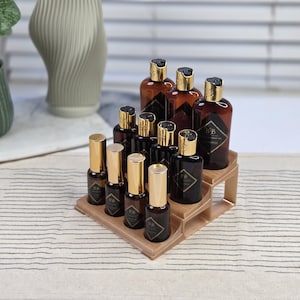 May include: A collection of brown glass bottles with gold lids and labels, displayed on a tiered wooden stand. The bottles are of different sizes and shapes, likely containing beauty or skincare products. The background features a vase and a window.