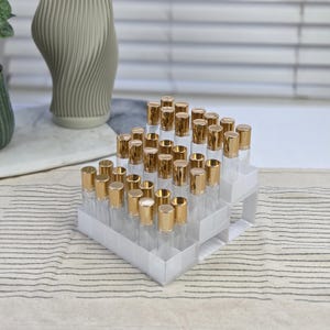 Könnte beinhalten: Ein mehrstufiger Ständer mit kleinen, klaren Glasflaschen mit goldfarbenen Kappen. Die Flaschen sind in Reihen auf einem weißen, mehrstufigen Organizer angeordnet. Der Hintergrund umfasst eine dekorative Vase und einen gestreiften Stoff.
