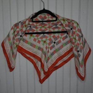 Peut inclure: Un foulard carré avec un motif géométrique blanc, orange, noir et vert. Le foulard est suspendu à un cintre noir contre un mur à panneaux blancs. Le foulard a une bordure orange.