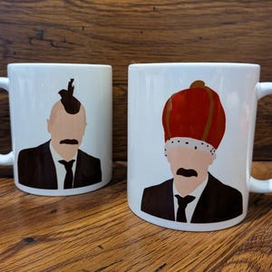 Könnte beinhalten: Zwei weiße Keramikbecher mit Illustrationen von Männern in Anzügen. Ein Becher zeigt einen Mann mit Irokesenschnitt, der andere einen Mann mit roter Krone. Beide Becher haben einen schwarzen Schnurrbart und eine Krawatte.