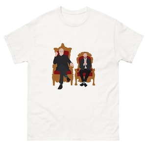 May include: A white short-sleeved t-shirt featuring a graphic illustration of two figures sitting in ornate brown chairs with red cushions. The figure on the left wears a black suit and glasses, while the figure on the right wears a dark jacket.