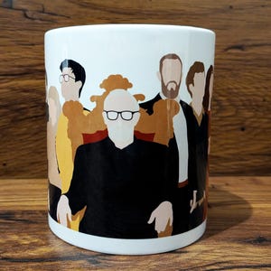 Può includere: Tazza in ceramica bianca con un disegno raffigurante un gruppo di figure stilizzate in nero, marrone e giallo. Le figure sono raffigurate con semplici contorni. La tazza ha una finitura lucida e una forma cilindrica.