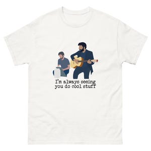 Könnte beinhalten: Weißes T-Shirt mit einer Illustration von zwei Personen, von denen eine Gitarre spielt und die andere an einem Laptop tippt. Der Text "I'm always seeing you do cool stuff" ist unter der Illustration aufgedruckt.