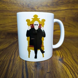 Könnte beinhalten: Weiße Keramiktasse mit einer Schwarzweißillustration einer Person mit Sonnenbrille, die auf einem goldenen Thron sitzt. Die Person macht mit beiden Händen eine Handgeste.