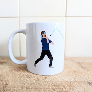 Könnte beinhalten: Weiße Keramik-Tasse mit einer Cartoon-Illustration eines Mannes, der einen Stein wirft. Der Mann trägt ein blaues Hemd, eine schwarze Hose und ein blaues Stirnband. Eine hellblaue Linie deutet die Flugbahn des Steins an.