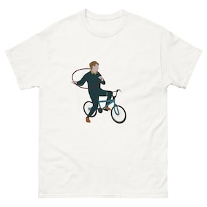 May include: White t-shirt with a graphic of a person on a blue bicycle, holding a hula hoop. The person wears a dark green outfit and brown shoes. The design is centered on the front of the shirt.