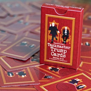 Peut inclure: Une boîte de jeu de cartes rouge avec le texte "Unofficial Taskmaster Trump Cards" et des images de deux personnages assis sur des chaises. Des cartes rouges similaires sont éparpillées.