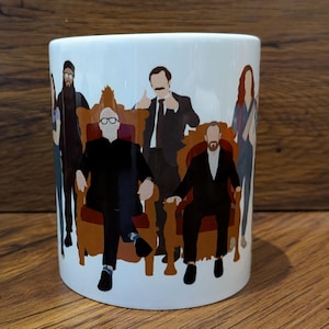 Könnte beinhalten: Weißer Keramikbecher mit einer stilisierten Illustration von mehreren Personen, von denen einige in verzierten Stühlen sitzen. Die Figuren sind in einem minimalistischen Stil mit Vollfarben und Umrissen dargestellt. Der Becher hat eine zylindrische Form.