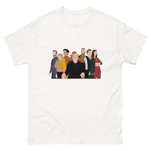 Puede incluir: Una camiseta blanca de manga corta con una ilustración gráfica central de ocho figuras estilizadas de pie juntas. Una figura central está sentada en una silla grande de color marrón, mientras que las demás están de pie en fila detrás.