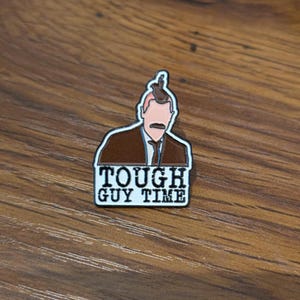 May include: A rectangular enamel pin featuring a cartoon image of a man in a brown suit with a mustache. The pin has the words "TOUGH GUY TIME" in white block letters on a black background.