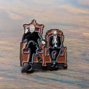May include: Enamel pin featuring two figures seated in ornate brown chairs. One figure wears a black suit and the other a black suit with a white shirt and tie. The pin has a detailed design with black, brown, and white colors.