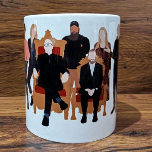 Puede incluir: Taza de cerámica blanca con un diseño de figuras estilizadas con trajes negros y ropa informal. Las figuras están dispuestas alrededor de un trono marrón. La taza es cilíndrica y descansa sobre una superficie de madera.