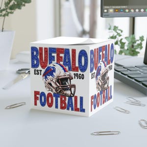 May include: A white cube notepad with the words "BUFFALO FOOTBALL" in red and blue, and a football helmet graphic. The text "EST 1960" is also included. The design is repeated on multiple sides of the cube.