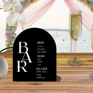May include: A black bar sign with white text listing beer, wine, and seltzer options. Two champagne flutes are filled with a bubbly beverage. A white orchid plant sits in a white pot. The background includes a beach scene and floral arch.