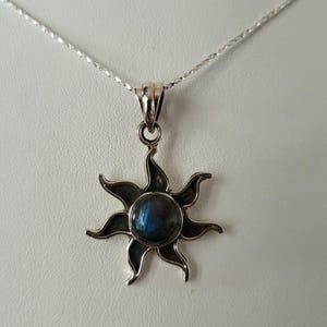 Könnte beinhalten: Halskette mit silbernem Sonnenanhänger. Der Anhänger hat einen dunkelblauen Edelstein in der Mitte, umgeben von stilisierten Strahlen. Die Halskette hat eine zarte Silberkette und einen einfachen Verschluss. Der Schmuck wird vor weißem Hintergrund präsentiert.