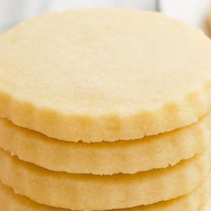 May include: A stack of four round, pale yellow shortbread cookies. Each cookie has a slightly scalloped edge and a smooth, lightly textured surface. The cookies are stacked neatly, showcasing their uniform shape and color.