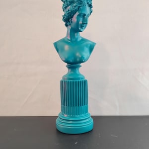 May include: A teal-colored bust of a woman with curly hair, mounted on a fluted pedestal. The sculpture is a solid teal color and has a classical design, suitable for home decor.