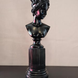 May include: A black bust sculpture of a woman with curly hair, mounted on a fluted pedestal base. The sculpture has a classical aesthetic and is made of a glossy, dark material. The bust is a decorative item.