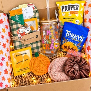 May include: A gift box containing a glass tumbler with a bamboo lid and straw featuring the text In My Pumpkin Spice Era, a bag of Proper Corn, a bag of Terry's Chocolate Orange Minis, a green plaid sleeve, eye masks, a crochet pumpkin, and a brown scrunchie.