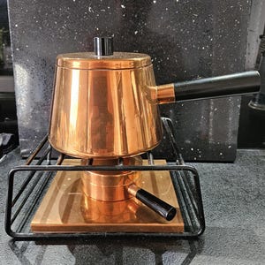 Puede incluir: Una olla de fondue de cobre con un mango negro se encuentra sobre un soporte de metal negro con una base de cobre. La olla tiene un pomo negro en la tapa.