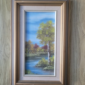 May include: A framed oil painting of a landscape scene with a single tree in the foreground. The tree is green and yellow, and the background is a blue sky with white clouds. The painting is signed "Ruddock" in the lower right corner.
