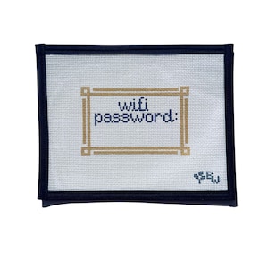 May include: A rectangular, white needlepoint with a navy blue border. The text "wifi password:" is centered within a gold-colored frame. A small logo is in the bottom right corner. The needlepoint is likely a decorative item.