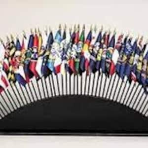 Puede incluir: Un soporte de banderas negro con un diseño en forma de abanico sostiene una colección de banderas en miniatura que representan varios países. Las banderas están dispuestas en semicírculo, con la base del soporte en la parte inferior.