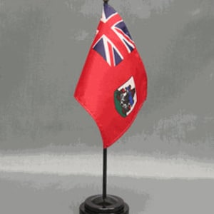 May include: A small red flag with the Union Jack and a coat of arms, mounted on a black stand with a gold finial. The flag is likely a miniature version of a national or regional flag, suitable for display on a desk or table.