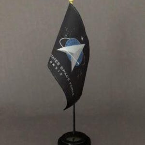 May include: A small black desk flag featuring the United States Space Force emblem. The flag has a white and blue design with the text "UNITED STATES SPACE FORCE MMXIX". The flag is mounted on a black stand with a gold finial.