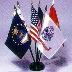 May include: A set of miniature desk flags featuring the United States flag, the United States Army flag with text, and other military-themed flags. The flags are mounted on thin black poles attached to a circular black base, arranged in a cluster.