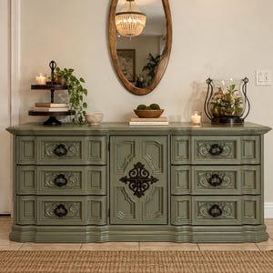 May include: A sage green wooden dresser featuring six drawers with ornate black metal pulls and a central cabinet door with a decorative black metal overlay. The piece has a rectangular top surface and sits on a base with molded trim details.
