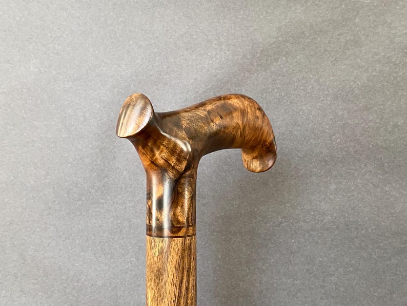 May include: A wooden walking stick with a curved handle. The handle and shaft are made of light brown wood with dark brown grain patterns. The cane is designed for support and mobility.