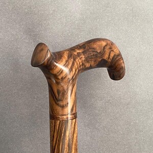 May include: A wooden walking cane with a curved handle. The cane is made of a dark brown wood with a unique grain pattern. The handle is smooth and rounded, providing a comfortable grip. The cane is a classic accessory.